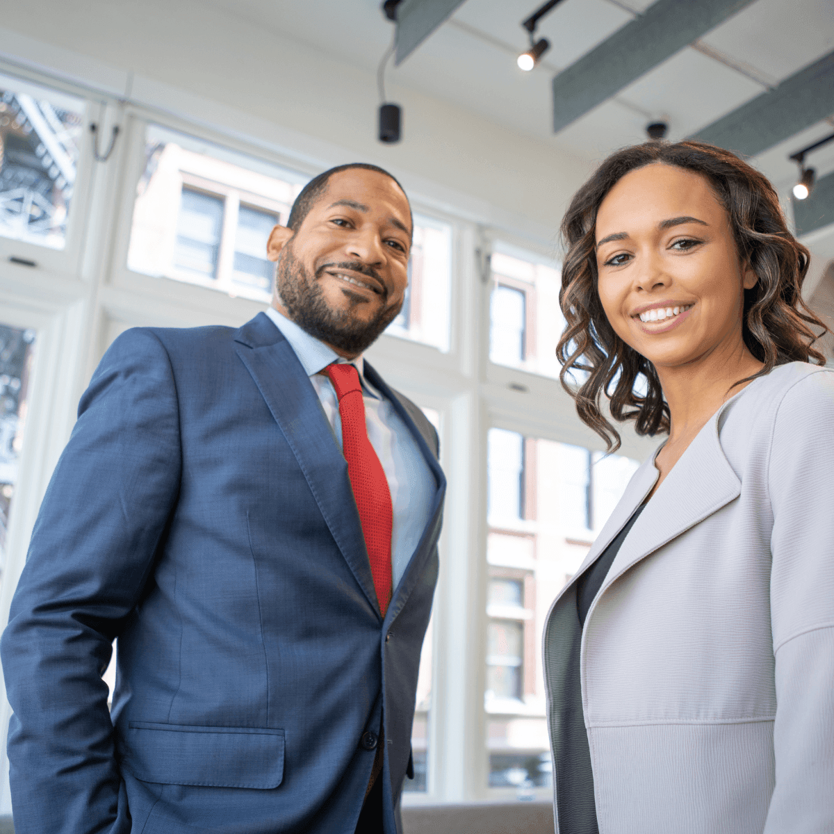 Two professionals in business attire standing in a well-lit office with large windows.