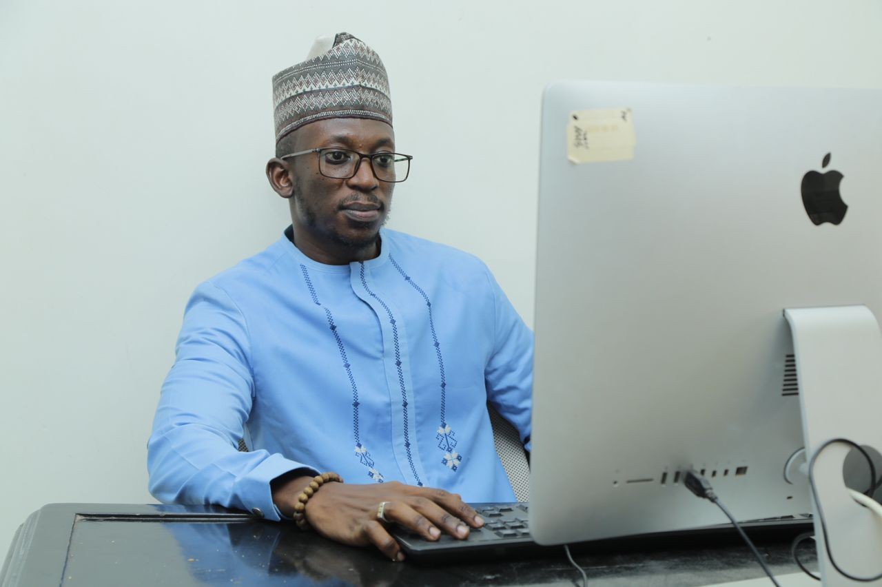 Man in blue traditional attire and cap working on an Apple computer.